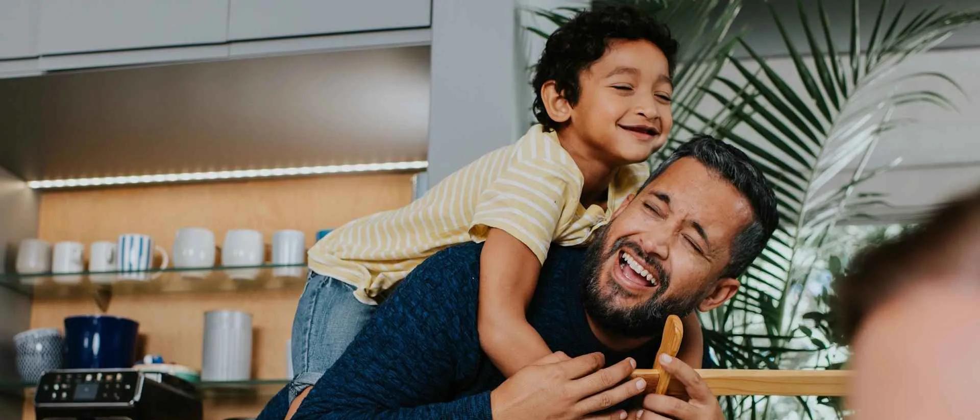 Padre e hijo felices comparten un paseo a caballito alegre y riendo en casa. Esta escena Philips destaca cómo nuestros productos, como la cafetera, apoyan el bienestar familiar.