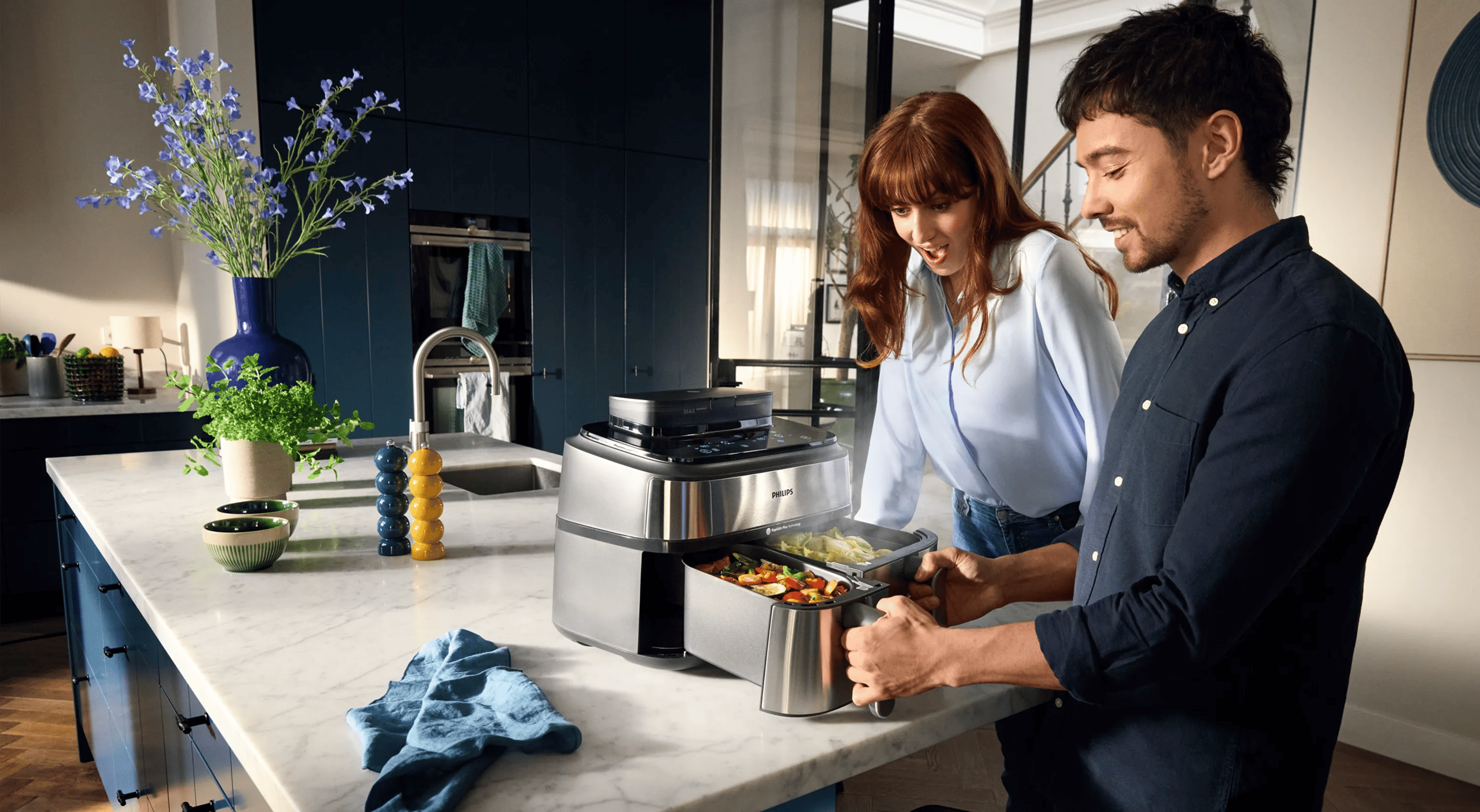 Paar genießt die Philips Kitchen Dual Basket Heißluftfritteuse, zeigt gefüllte Körbe mit Gemüse, Dampf steigt auf. Moderne Küche mit blauen Schränken und Marmorarbeitsplatte.