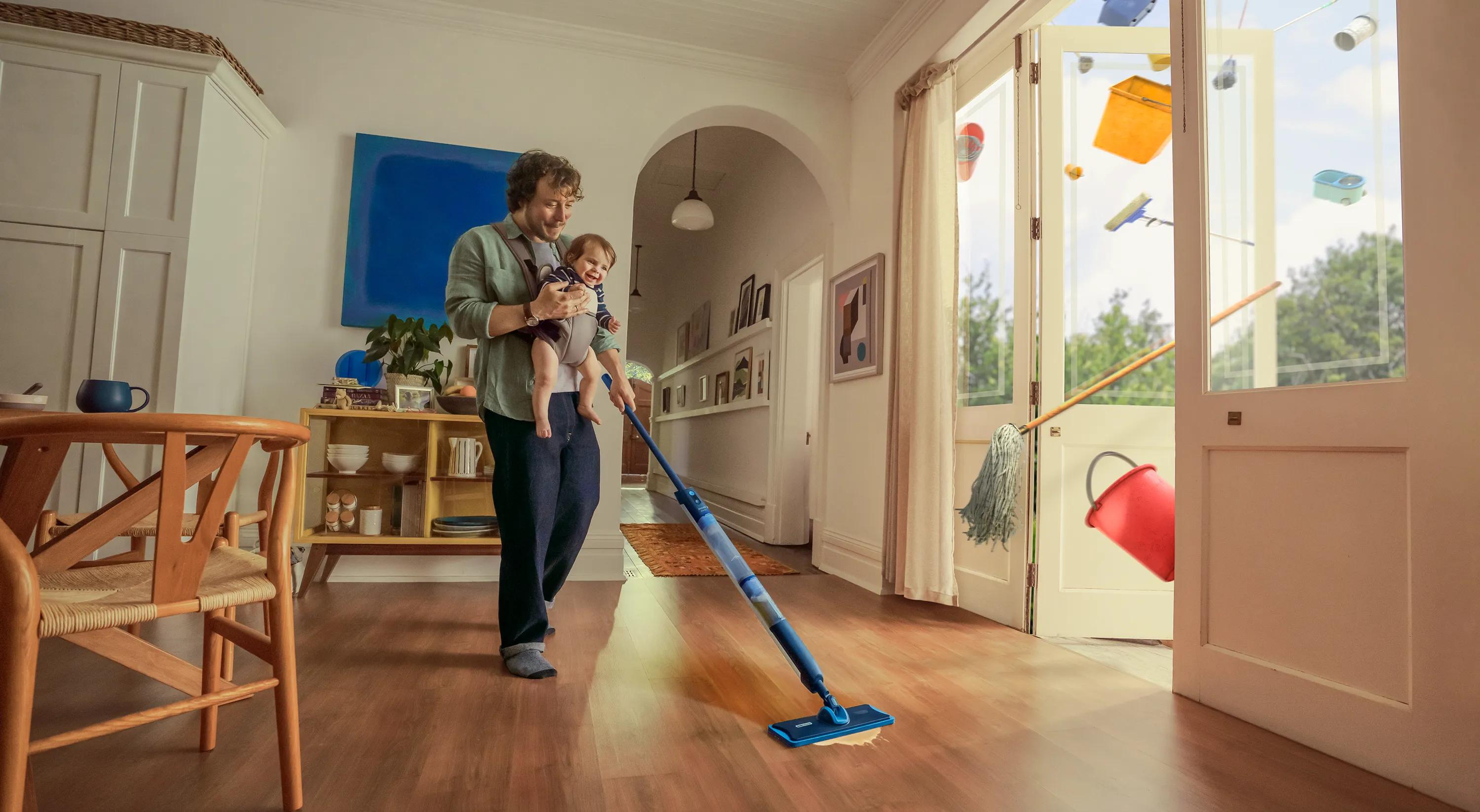 Člověk vytírá podlahu v jasném moderním obývacím pokoji, zatímco drží dítě v náručí; barevné předměty z domácnosti se vznáší ven skrz otevřené skleněné dveře.