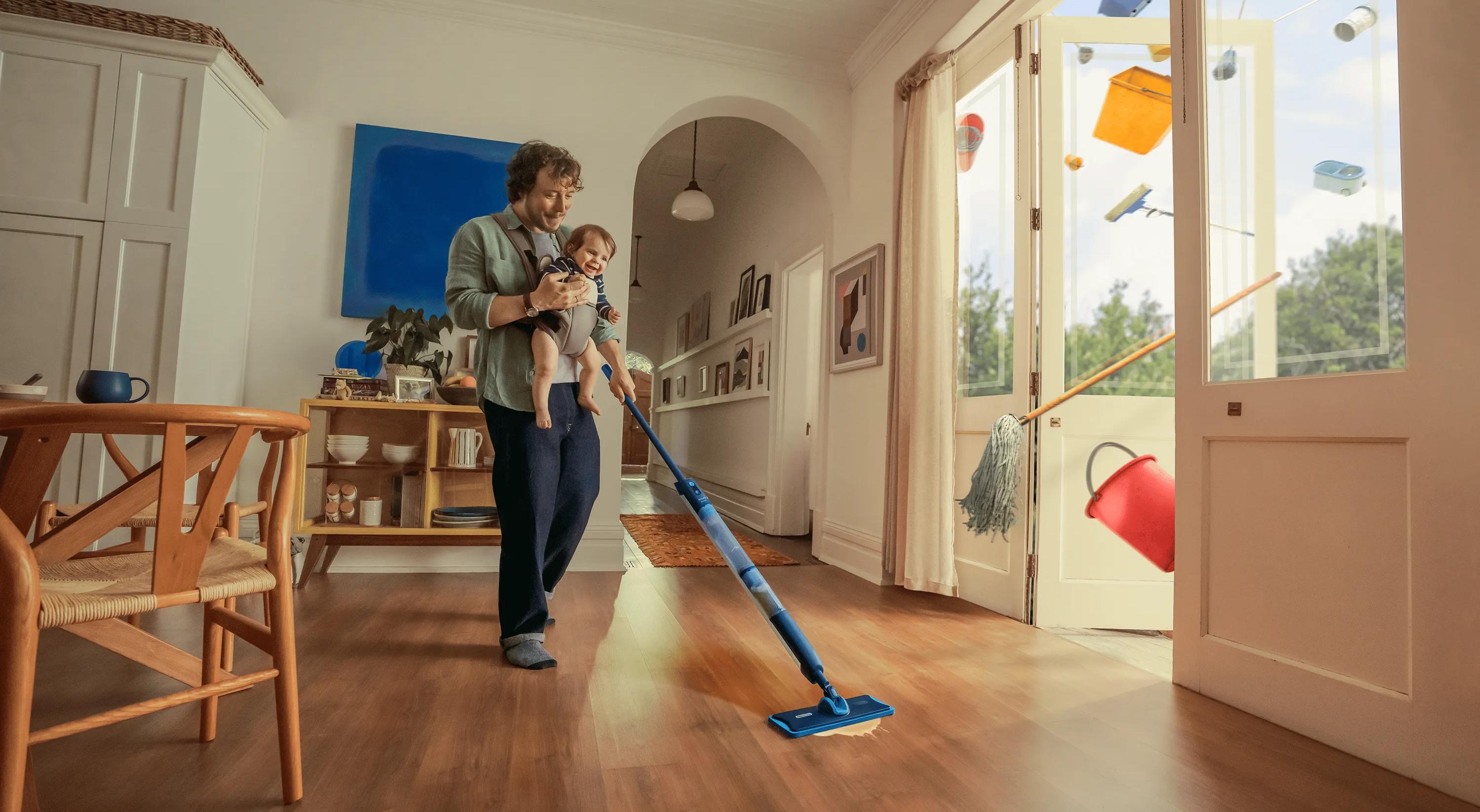 Eine Person wischt ein helles, modernes Wohnzimmer, während sie ein Baby im Arm hält; bunte Haushaltsgegenstände schweben durch die offenen Glastüren nach draußen.