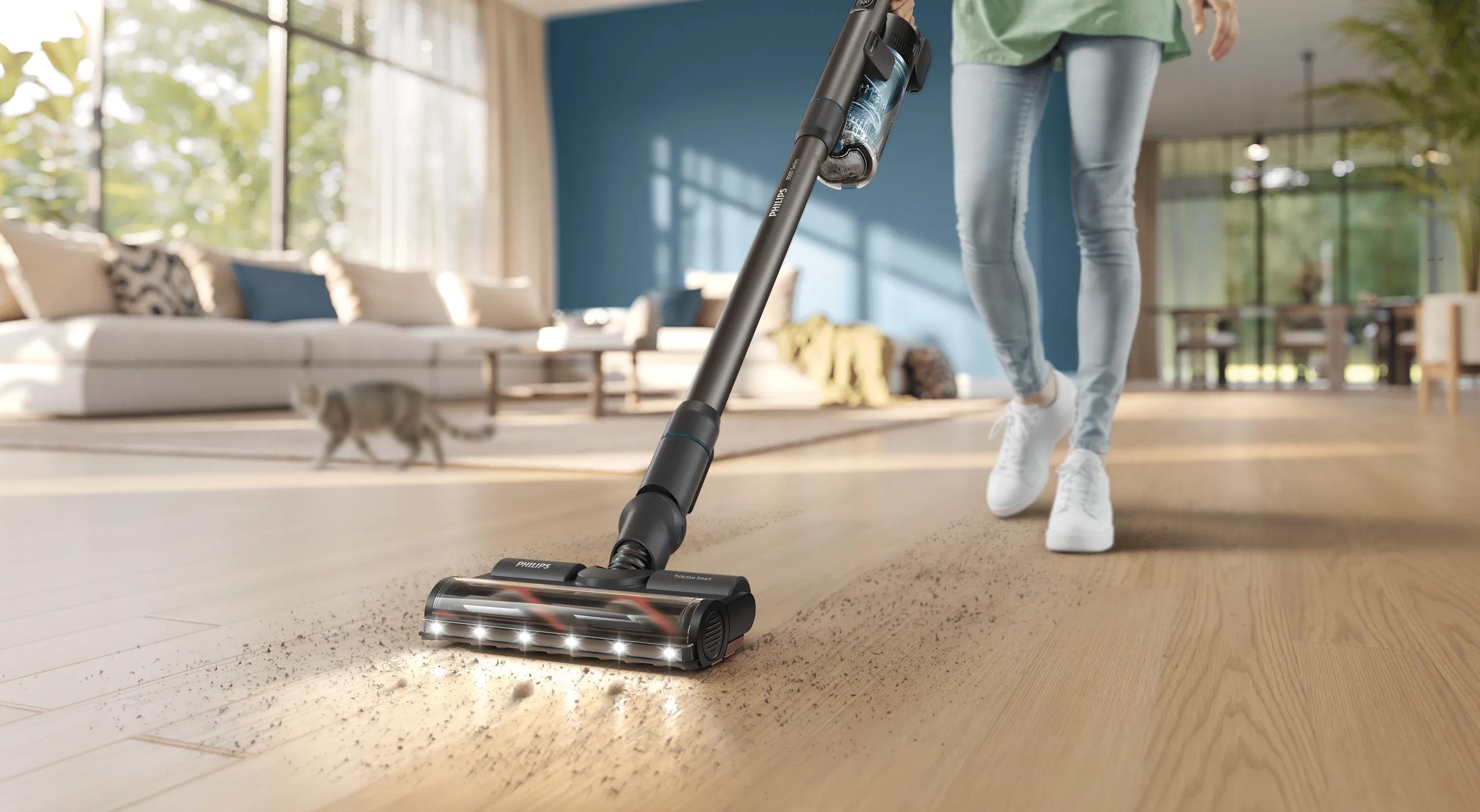 Persona limpiando un suelo de madera con una aspiradora escoba Philips en un luminoso salón moderno con un gato en el fondo