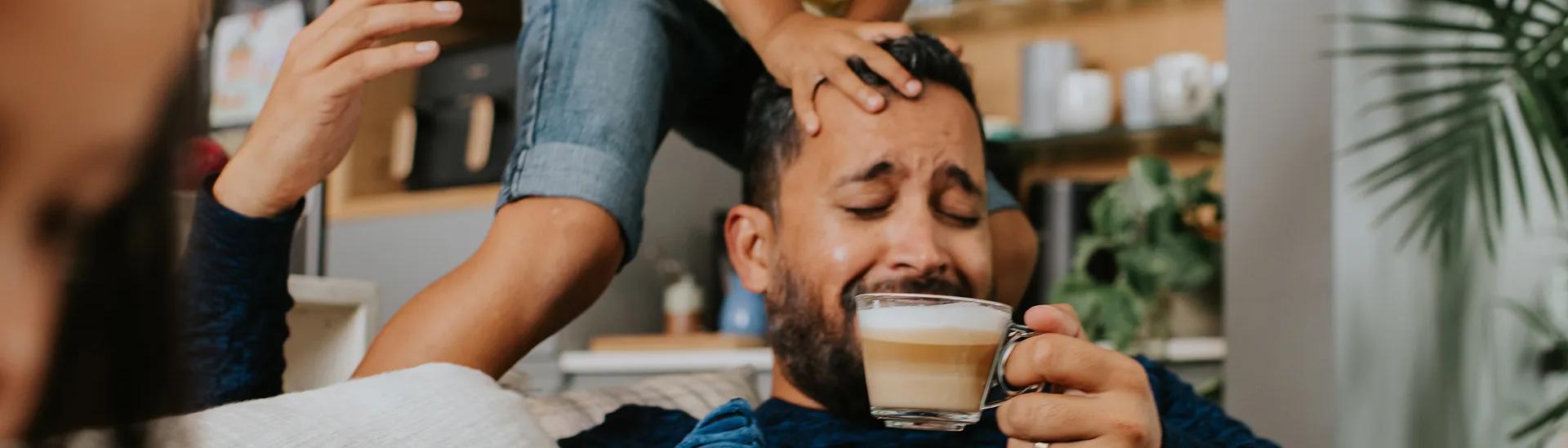 Mann lächelt beim Kaffeetrinken zu Hause, während sein Kind über seinen Kopf springt