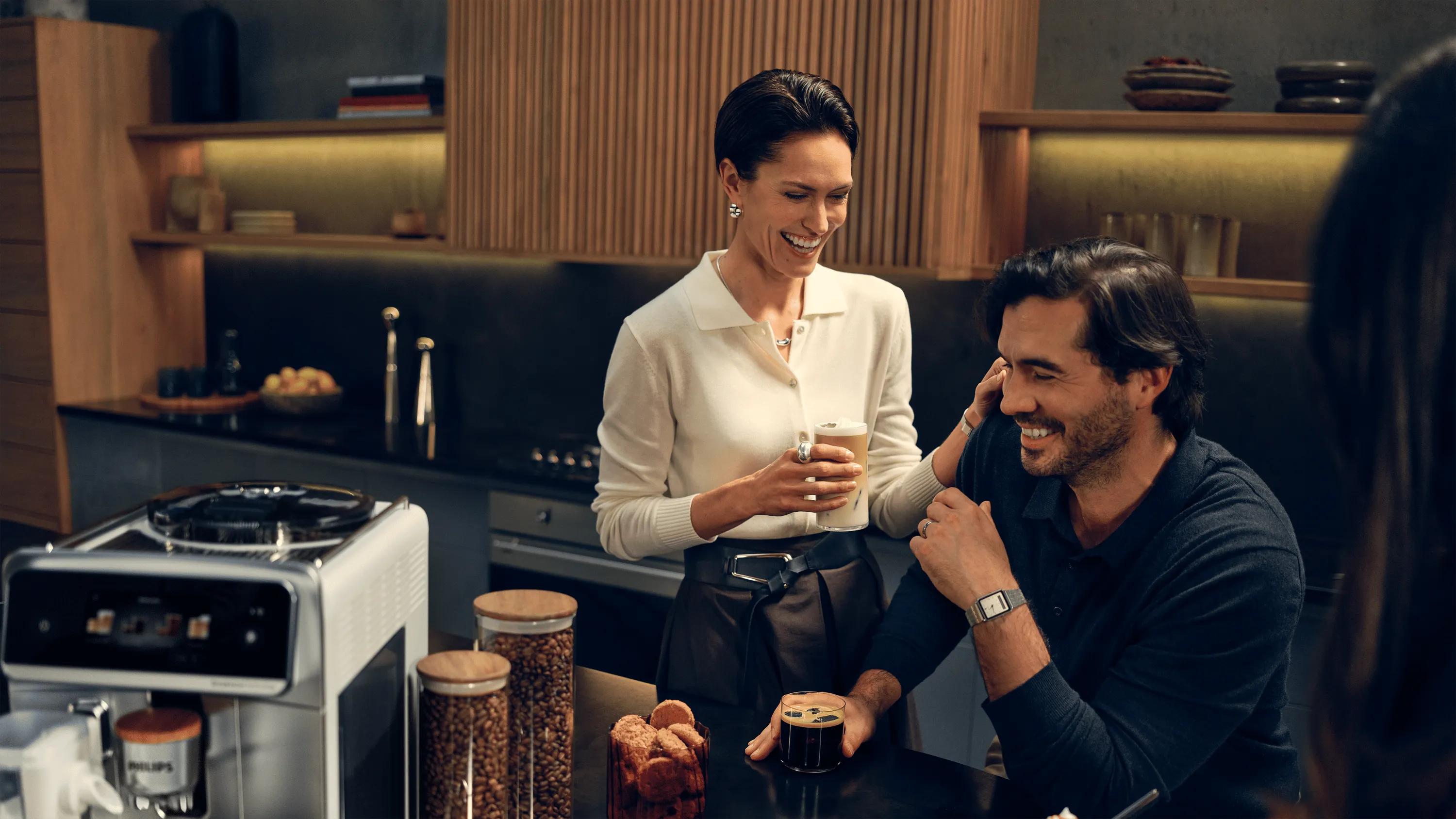 Kávovar Philips Café Aromis řady 8000 na moderní kuchyňské lince, zatímco pár si doma vychutnává čerstvě připravenou kávu