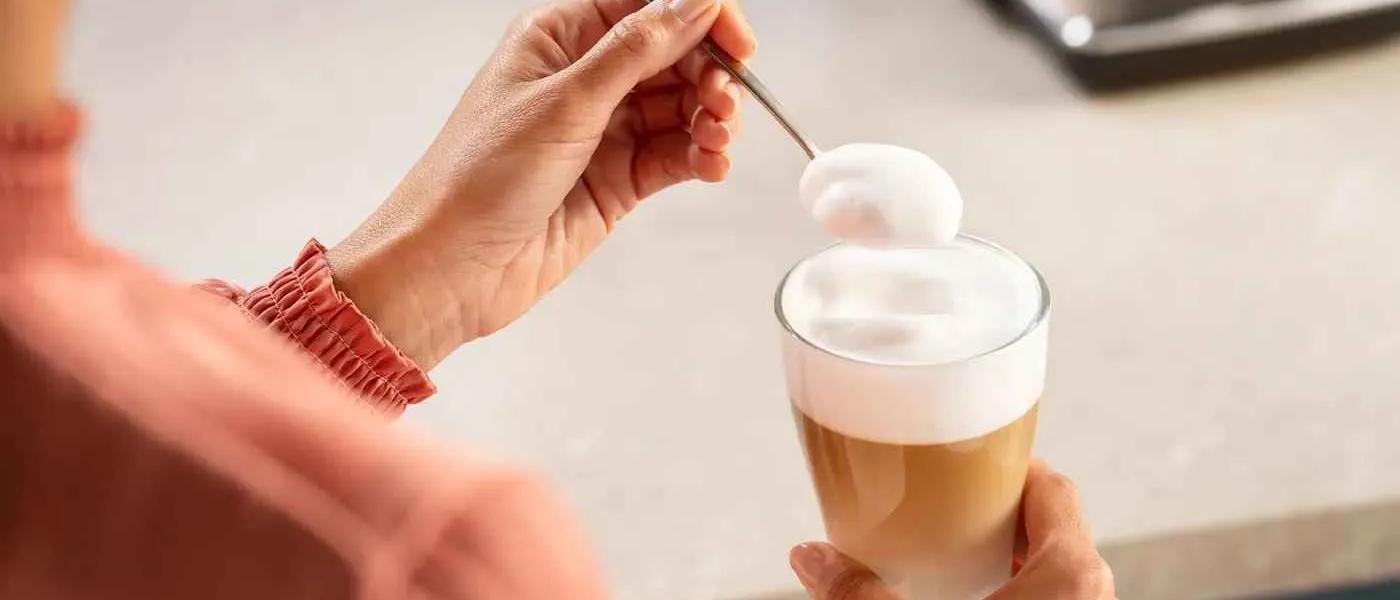 En person tilføjer tæt mælkeskum med en ske til en lagdelt Philips LatteGo latte, hvilket fremhæver perfekt skum til cafékvalitetsdrikke hjemme.