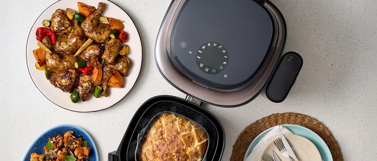 Philips Airfryer 5000 mit geöffnetem Korb, der einen gekochten Kuchen im Inneren zeigt. Um den Airfryer herum befinden sich Teller mit gekochten Hähnchenflügeln und geröstetem Gemüse, Besteck auf einer Serviette und eine moderne Küchenarbeitsplatte.